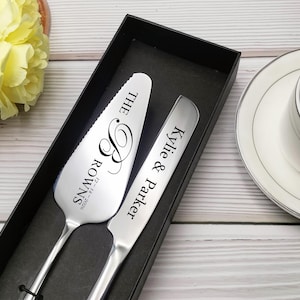 Peut inclure: Un ensemble de serveur et de couteau à gâteau argentés dans une boîte noire. Le serveur est gravé de "THE ROWNS" et d'une date. Le couteau est gravé de "Kylie & Parker". Une tasse de café est posée sur une soucoupe en arrière-plan.