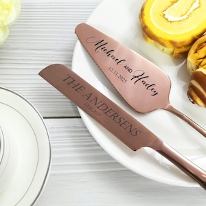 Pode incluir: Conjunto de faca e espátula para bolo em ouro rosa com gravação personalizada. A espátula está inscrita com "Michael and Hailey 12.11.2025". A faca está inscrita com "THE ANDERSENS 12.11.2025". O conjunto está sobre um prato branco com doces.