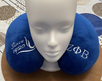 May include: A royal blue U-shaped travel neck pillow featuring white embroidered text. The left side displays the words "Since 1920" with a swoosh design, while the right side features the Greek letters "ZΦB" in a clean, white, stitched font.