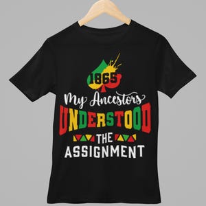 May include: Black t-shirt with the text "1865 My Ancestors Understood The Assignment." The design features a colorful graphic with red, yellow, and green accents. The shirt is hanging on a wooden hanger.