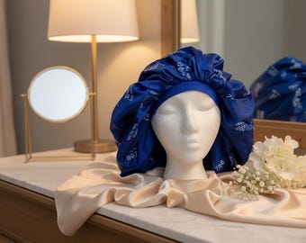 May include: A royal blue satin bonnet featuring a repeating white Greek letter pattern and a solid blue elasticized band. The bonnet is displayed on a white mannequin head, showcasing the gathered fabric design and the printed text.