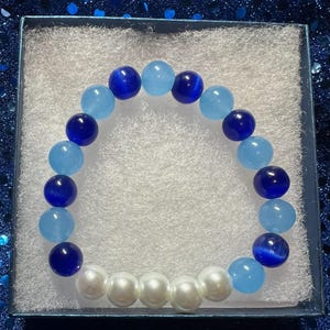 Puede incluir: Una pulsera de cuentas con cuentas esféricas azul claro y azul oscuro alternadas, y una fila de perlas blancas. La pulsera se presenta en una caja de regalo azul con acolchado blanco. Las cuentas miden aproximadamente 1,3 cm de diámetro.