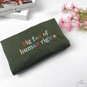 May include: An olive green sweatshirt with the embroidered text "big fan of human rights" in colorful letters. The folded garment is on a white surface, with a book and pink flowers in the background.