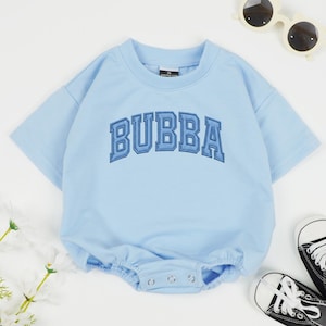 May include: A light blue baby romper with the word "BUBBA" in dark blue block letters. The romper has short sleeves and snaps at the bottom. A pair of black and white baby sneakers and white sunglasses are also in the image.