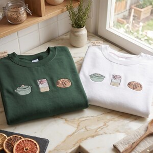 May include: Two sweatshirts, one dark green and one white, each with embroidered kitchen-themed designs. The green sweatshirt features a pot, jar, and bread. The white sweatshirt has the same design. The text "Cozy Kitchen Collection" is visible.