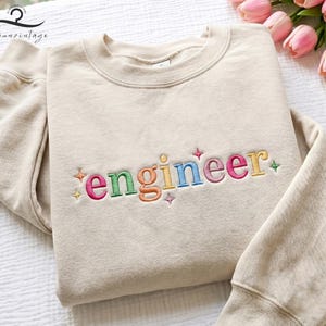 May include: A beige sweatshirt with the word "engineer" embroidered in colorful letters. The word is in a rainbow of colors, with star accents. The sweatshirt is folded on a white surface, with a bouquet of pink tulips in the background.