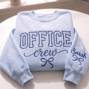 May include: A light blue sweatshirt with the words "OFFICE crew" embroidered in dark blue. The word "OFFICE" has a gingham pattern. The right sleeve has the name "Sarah" embroidered. A bow design is below the word "crew".