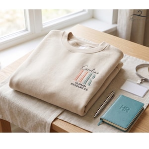 May include: Beige crewneck sweatshirt with embroidered "Caroline HR HUMAN RESOURCES" design in black, pink, and teal. A teal notebook with "HR" embossed, a pen, and a name tag are on a wooden table.