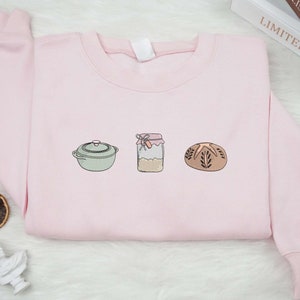 May include: A pink sweatshirt with a graphic of a pot, a jar, and a loaf of bread. The graphic is in a light green, brown, and white color scheme.