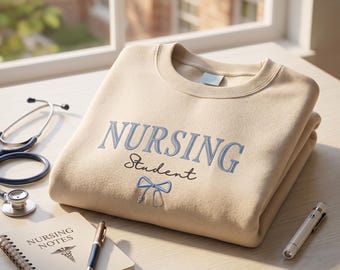 May include: A beige sweatshirt with the words "NURSING Student" embroidered in light blue. A stethoscope, notebook, pen, and penlight are arranged around the sweatshirt on a wooden surface. The notebook reads "NURSING NOTES".