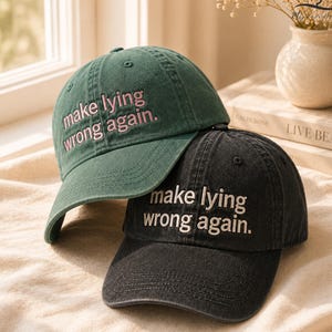 May include: Two baseball caps, one dark green and one black, are stacked. The green cap has pink embroidered text that reads "make lying wrong again." The black cap has white embroidered text that reads the same.