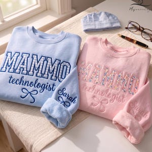 May include: A light blue sweatshirt and a light pink sweatshirt, both featuring the embroidered text MAMMO technologist with a bow icon and the name Sarah on the sleeve. The blue sweatshirt has a gingham pattern, while the pink has a floral pattern.