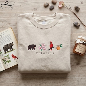 May include: A cream-coloured sweatshirt featuring embroidered Virginia state symbols: a bear, dogwood flowers, a cardinal, and a peach. The word "VIRGINIA" is embroidered below. A jar of Virginia Peach Preserves is in the background.