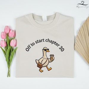 May include: Beige t-shirt with the embroidered phrase "Off to start chapter 30." The shirt features a cartoon goose wearing sunglasses, a pearl necklace, and a purse, holding a coffee cup. The design is centered on the front of the shirt.