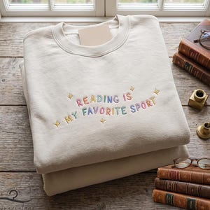 May include: A cream-colored crewneck sweatshirt featuring the embroidered text "READING IS MY FAVORITE SPORT" in multi-colored letters, accented with small embroidered star shapes. The garment is folded and displayed on a wooden surface.