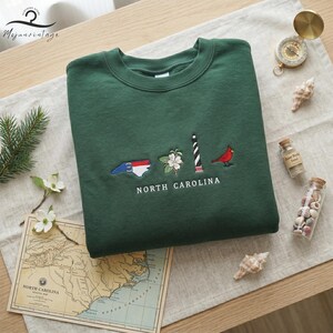 K&ouml;nnte beinhalten: Dunkelgr&uuml;nes Sweatshirt mit Stickereien der Symbole des US-Bundesstaates North Carolina: Flagge, Blume, Leuchtturm und Kardinal. Der Text "NORTH CAROLINA" ist darunter gestickt. Eine Karte und dekorative Gegenst&auml;nde umgeben das Sweatshirt.