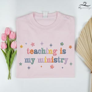 May include: A light pink t-shirt with the embroidered text "teaching is my ministry" in colorful letters. The shirt is decorated with embroidered flowers and stars. A bouquet of pink tulips and a decorative fan are in the background.