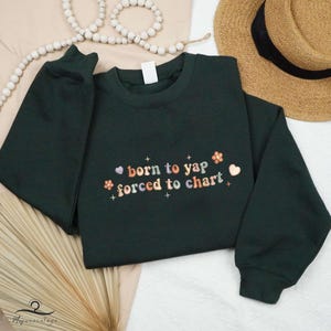 May include: A dark green sweatshirt with the text "born to yap forced to chart" in colorful letters with small flower and star accents.