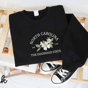 May include: Black sweatshirt with a white floral design and the text "North Carolina" above "The Dogwood State." The design includes the year 1789. A pair of black and white sneakers are next to the sweatshirt.