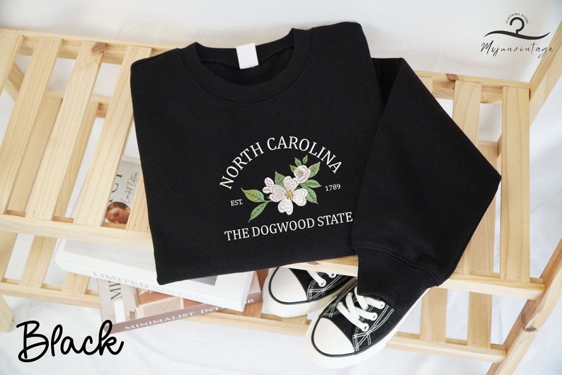 May include: Black sweatshirt with a white floral design and the text "North Carolina" above "The Dogwood State." The design includes the year 1789. A pair of black and white sneakers are next to the sweatshirt.