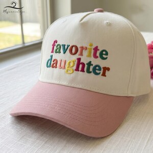 Puede incluir: Una gorra de b&eacute;isbol de color crema con visera rosa. El panel frontal presenta el texto bordado "favorite daughter" en letras multicolores, incluyendo tonos de rosa, verde, rojo, amarillo y azul, con un bot&oacute;n en la parte superior.