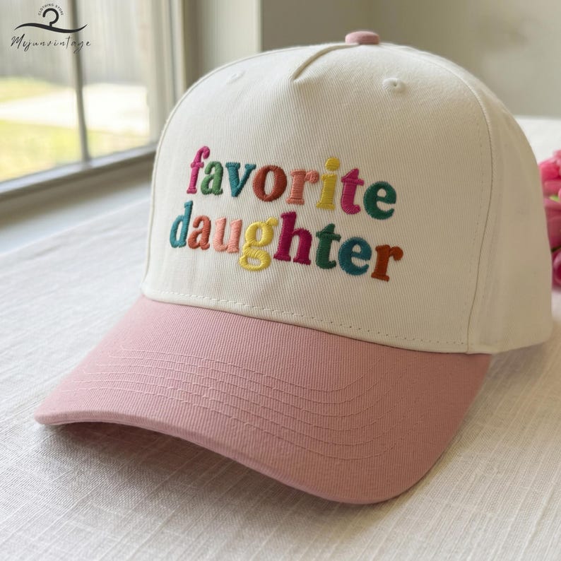 Puede incluir: Una gorra de b&eacute;isbol de color crema con visera rosa. El panel frontal presenta el texto bordado "favorite daughter" en letras multicolores, incluyendo tonos de rosa, verde, rojo, amarillo y azul, con un bot&oacute;n en la parte superior.