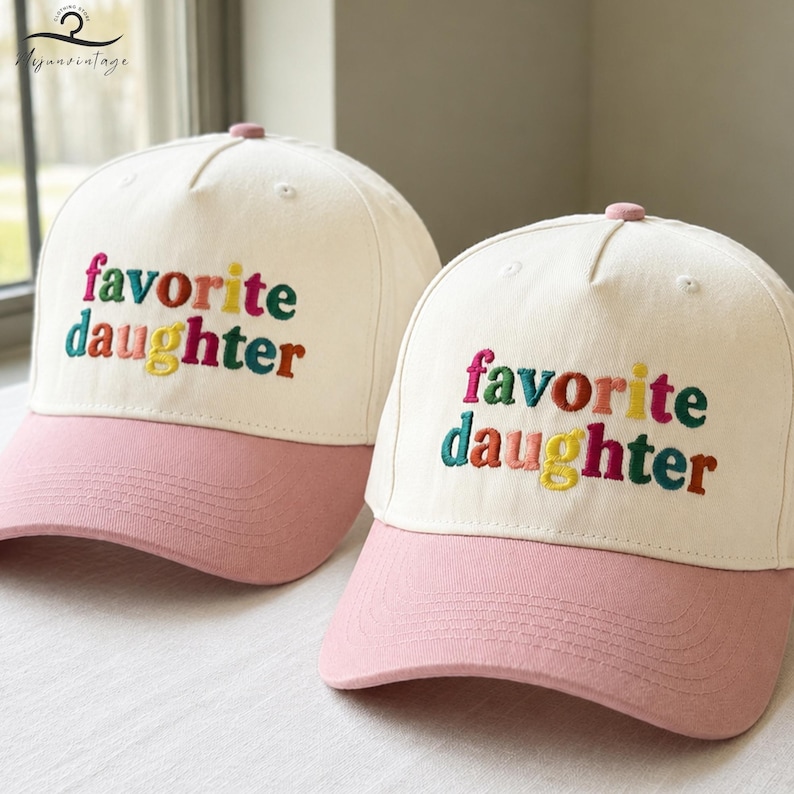 Puede incluir: Dos gorras de b&eacute;isbol con corona color blanco hueso y visera rosa. Cada gorra presenta el texto bordado "favorite daughter" en la parte frontal con letras multicolores, incluyendo tonos rosa, rojo, amarillo y azul verdoso.