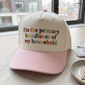 May include: A baseball cap with a cream-colored crown and a pink brim. The cap features embroidered text in various colors that reads, "I'm the primary breadloser of my household."