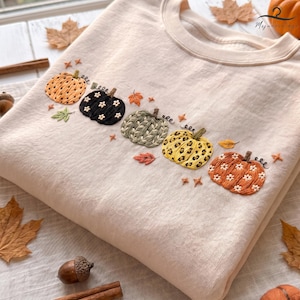 May include: A beige crewneck sweatshirt featuring an embroidered horizontal row of five pumpkins. The pumpkins display varied patterns including orange with white flowers, black with white flowers, green leaves, yellow leopard print, and orange with white flowers.