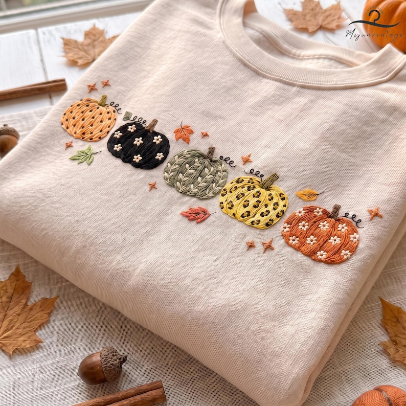 May include: A beige crewneck sweatshirt featuring an embroidered horizontal row of five pumpkins. The pumpkins display varied patterns including orange with white flowers, black with white flowers, green leaves, yellow leopard print, and orange with white flowers.