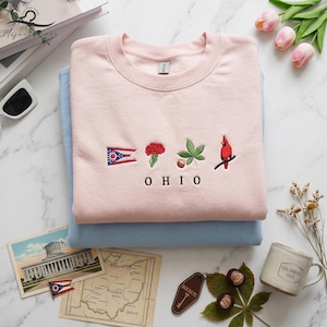 May include: A stack of two sweatshirts, one light pink and one light blue. The pink sweatshirt has embroidered Ohio state symbols: a flag, a red flower, a leaf, and a cardinal. The word "OHIO" is embroidered below the symbols.