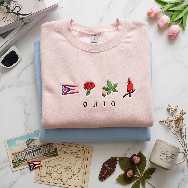 May include: A stack of two sweatshirts, one light pink and one light blue. The pink sweatshirt has embroidered Ohio state symbols: a flag, a red flower, a leaf, and a cardinal. The word "OHIO" is embroidered below the symbols.