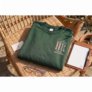 May include: A dark green crew neck sweatshirt with the text "Caroline HR Human Resources" in a script font and a striped design. The sweatshirt is on a wicker chair next to a notepad and glasses.