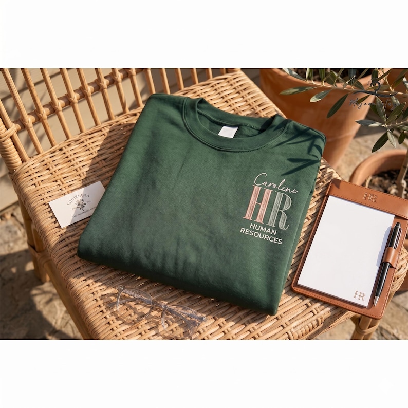 May include: A dark green crew neck sweatshirt with the text "Caroline HR Human Resources" in a script font and a striped design. The sweatshirt is on a wicker chair next to a notepad and glasses.