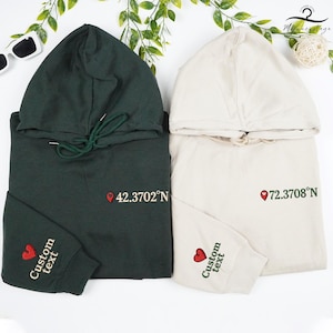 May include: Two hooded sweatshirts, one dark green and one beige. Each features a location pin graphic with coordinates and the text "Custom text" on the sleeve. The green hoodie has a green drawstring, and both have a heart graphic.
