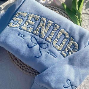 May include: Light blue sweatshirt with a floral embroidered "Senior" design and the text "EST. 2026".