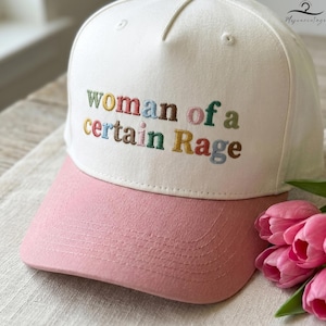 May include: A white baseball cap with a pink bill. The cap features the embroidered phrase "woman of a certain Rage" in colorful letters. A bouquet of pink tulips is in the background.