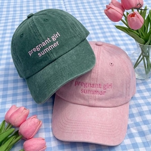 K&ouml;nnte beinhalten: Zwei Baseballkappen, eine gr&uuml;ne und eine rosa, werden gezeigt. Auf den Kappen sind die Worte "pregnant girl summer" aufgestickt. Die Kappen liegen auf einer blau-wei&szlig; karierten Oberfl&auml;che, mit rosa Tulpen im Hintergrund.