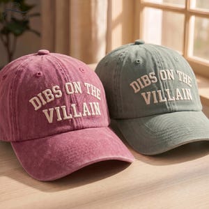 May include: Two baseball caps, one in a faded berry red and one in a muted sage green, both featuring the embroidered white text DIBS ON THE VILLAIN across the front panels. Each cap has a structured crown, a curved brim, and eyelet details.