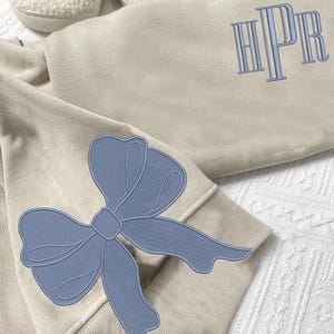 May include: A beige sweatshirt with a blue bow on the sleeve and the monogram "HPR" embroidered on the front.