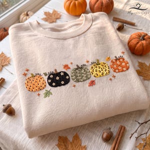 May include: A cream-colored crewneck sweatshirt featuring an embroidered row of five pumpkins in various colors and patterns, including orange, black, green, yellow, and rust, accented with small white flowers and scattered autumn leaves.