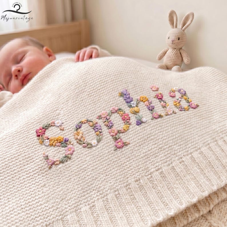 May include: A cream-coloured knit baby blanket featuring the name Sophia embroidered in a floral pattern. The letters are composed of small, colourful embroidered flowers in shades of pink, yellow, purple, and green. A small crocheted rabbit toy sits nearby.