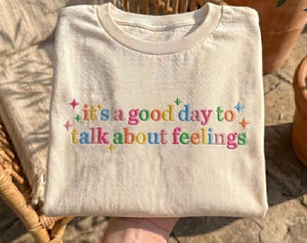 May include: Cream-colored t-shirt with the embroidered text "it's a good day to talk about feelings" in colorful thread. The shirt is folded and held by a person. The design includes star accents.