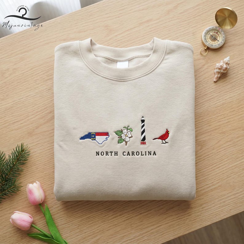 K&ouml;nnte beinhalten: Beiges Sweatshirt mit gestickten Symbolen von North Carolina. Das Design umfasst eine Kartenumriss, eine Kornblume, einen Leuchtturm und einen roten Kardinal. Der Text "NORTH CAROLINA" ist darunter gestickt.