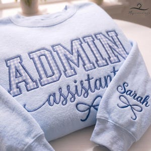 May include: Light blue sweatshirt with embroidered text. The word "ADMIN" is in large, block letters with a blue and white gingham pattern. The word "assistant" is in cursive, and the name "Sarah" is on the sleeve.