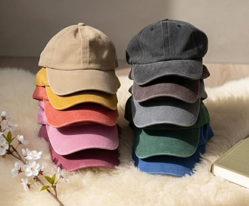 K&ouml;nnte beinhalten: Gestapelte Baseballkappen in verschiedenen Farben: links beige, gelb, orange, rosa und weinrot, rechts grau, braun, gr&uuml;n und blau. Die Kappen liegen auf einer flauschigen, wei&szlig;en Oberfl&auml;che.