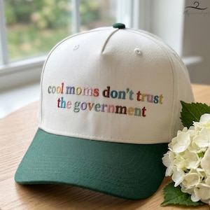 May include: A white baseball cap with a green brim and top button. The cap features embroidered text in various colors that reads "cool moms don't trust the government". The cap is sitting on a wooden surface.