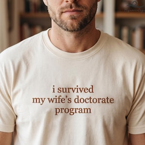 May include: Cream-colored t-shirt with the text "i survived my wife's doctorate program" in brown lettering. The shirt has a crew neck and short sleeves. The image is a close-up of the shirt on a person.