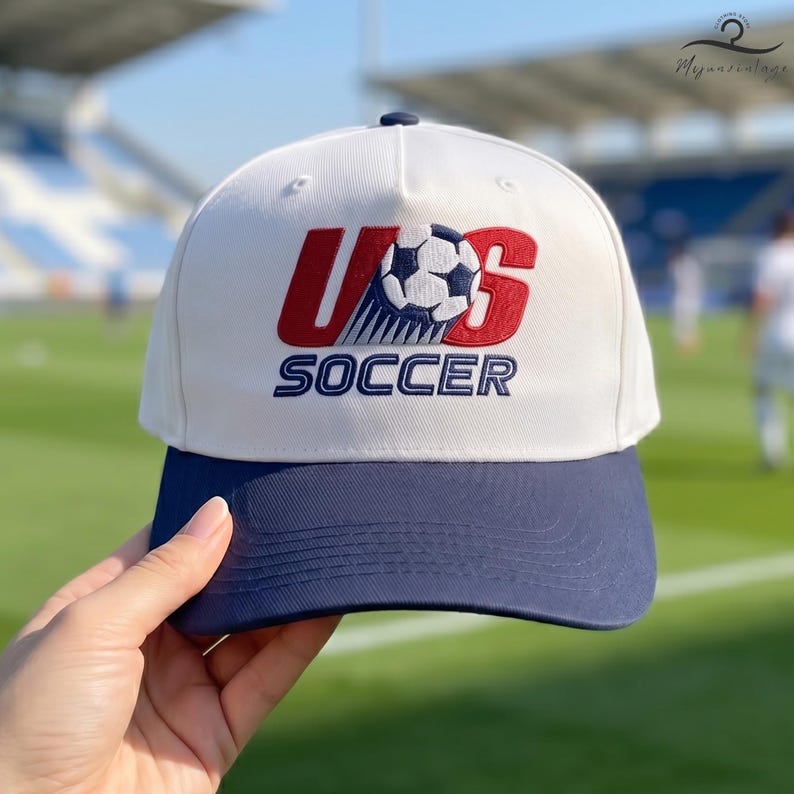 May include: A white baseball cap with a navy blue brim. The front features an embroidered logo with the red letters U and S flanking a black and white football graphic, with the word SOCCER embroidered in navy blue text underneath the graphic.