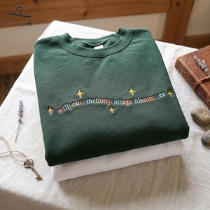 May include: A dark green sweatshirt with the embroidered text "willyoucometomycottage hissummer" in rainbow colors, accented with gold stars. The sweatshirt is folded on top of a white garment. A key and lavender sprig are visible.